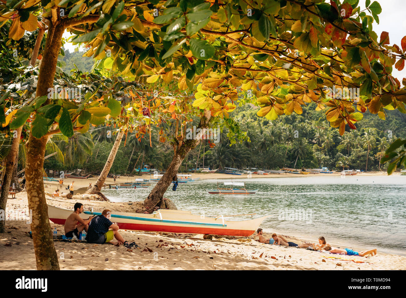 Port Barton, Palawan, Philippines - February 3, 2019: People relax on tropical beach under the lush crown of trees Stock Photo
