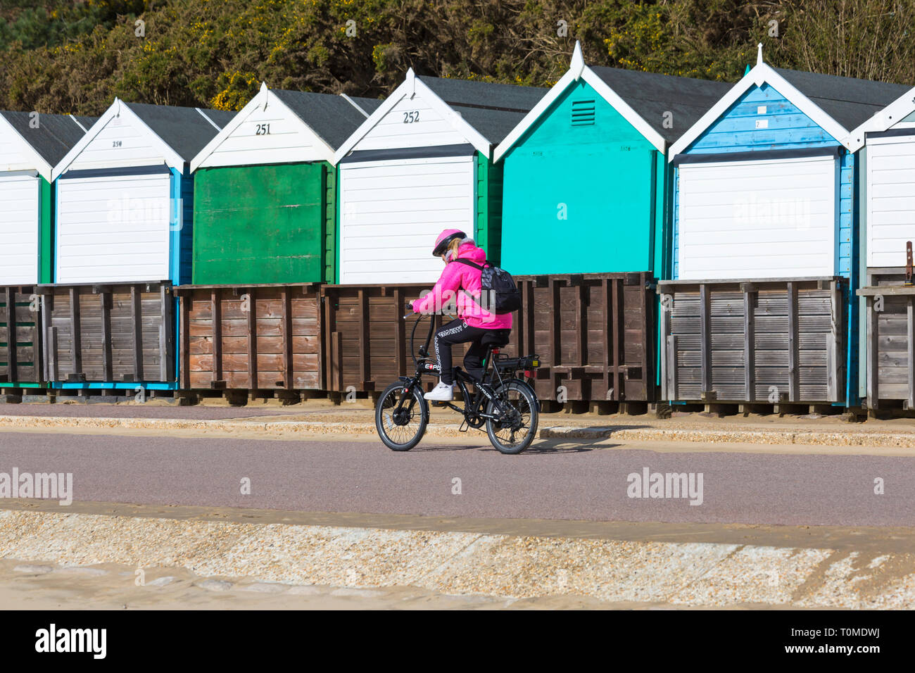 Young woman cycling, riding an eco power assisted electric bike bicycle along promenade past beach huts at Bournemouth, Dorset UK in March Stock Photo