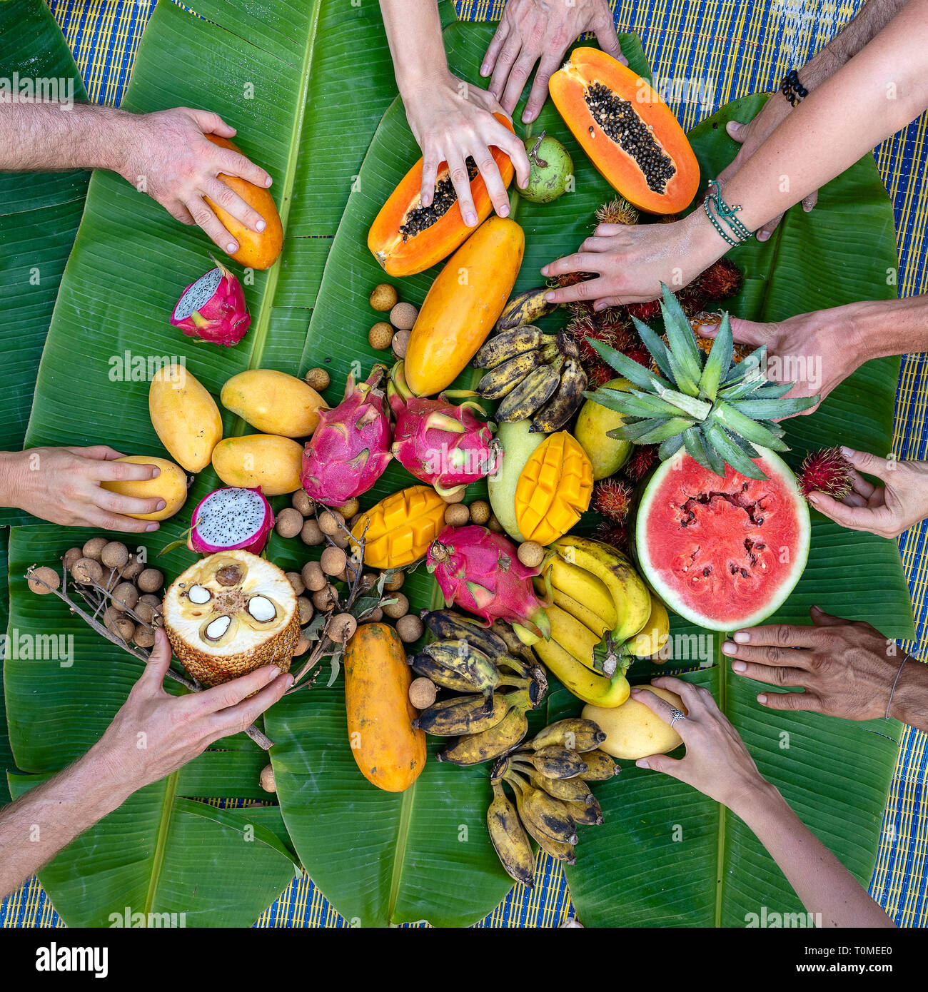 Tropical fruits on green banana leaves and people hands. Group of happy friends having nice food, enjoying the party and communication. Mango, papaya, Stock Photo