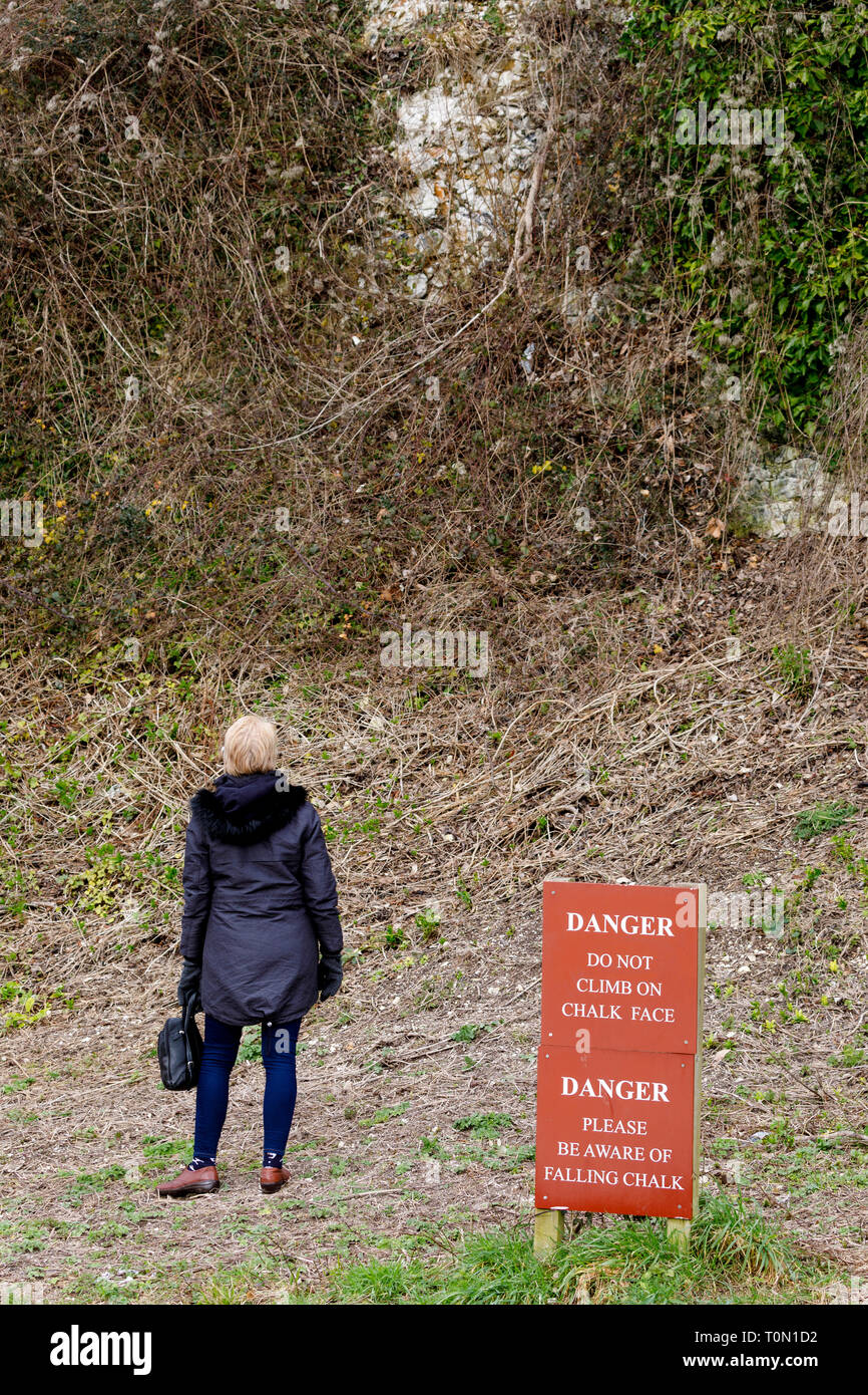 Woman stands beside a warning sign at the bottom of a chalk escarpment in Sussex, UK. Stock Photo