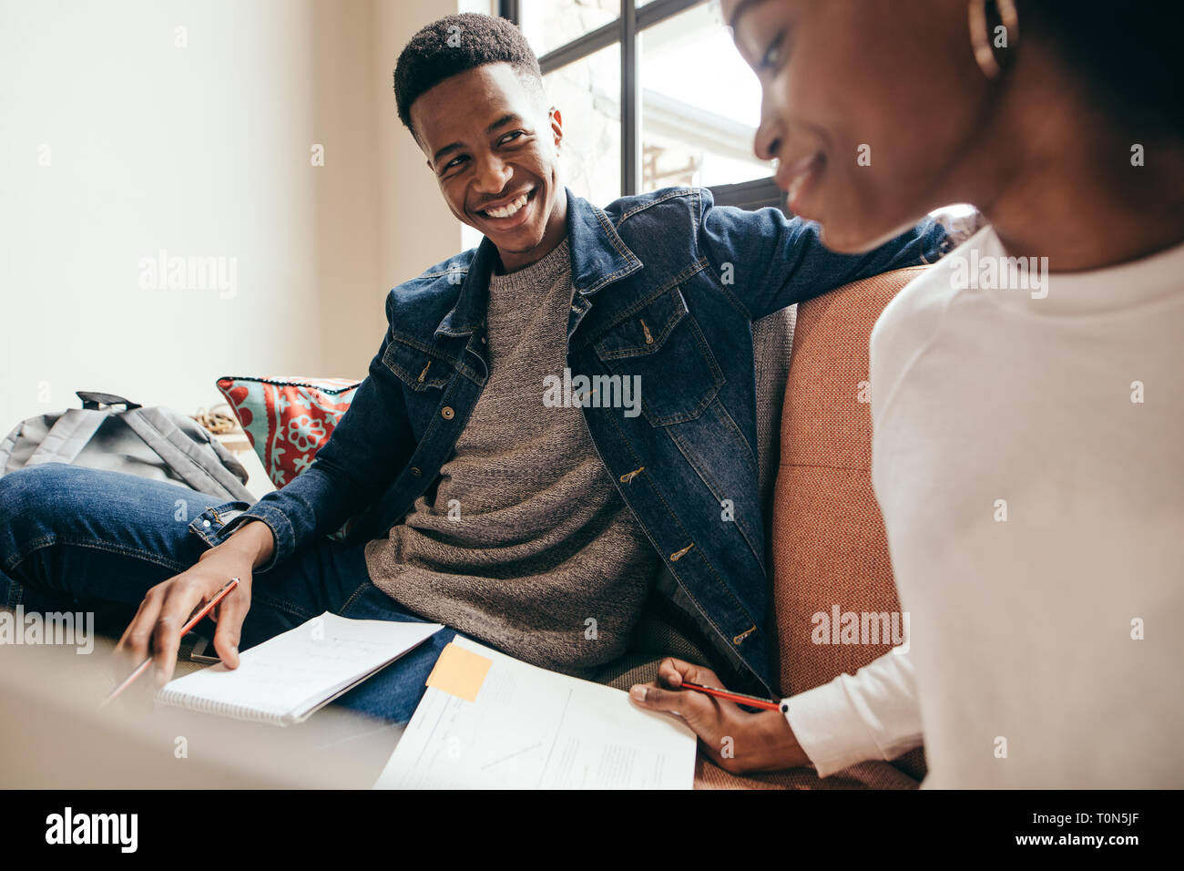 Smiling teenager looking at girl reading book on sofa at university