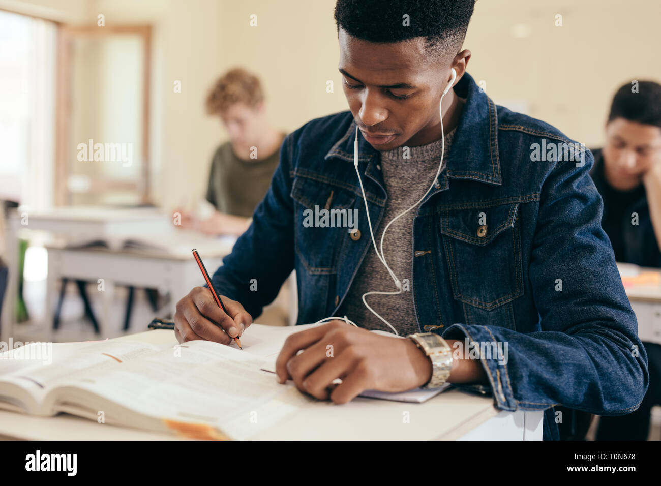 Teenage boy sitting in high school classroom writing in book. Male student with earphones on, making notes during lecture in college classroom. Stock Photo