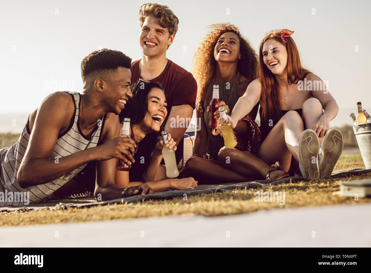 Group of diverse young people relaxing outdoors with drinks. Young men and women hanging out, chatting, having beers and laughing. Stock Photo