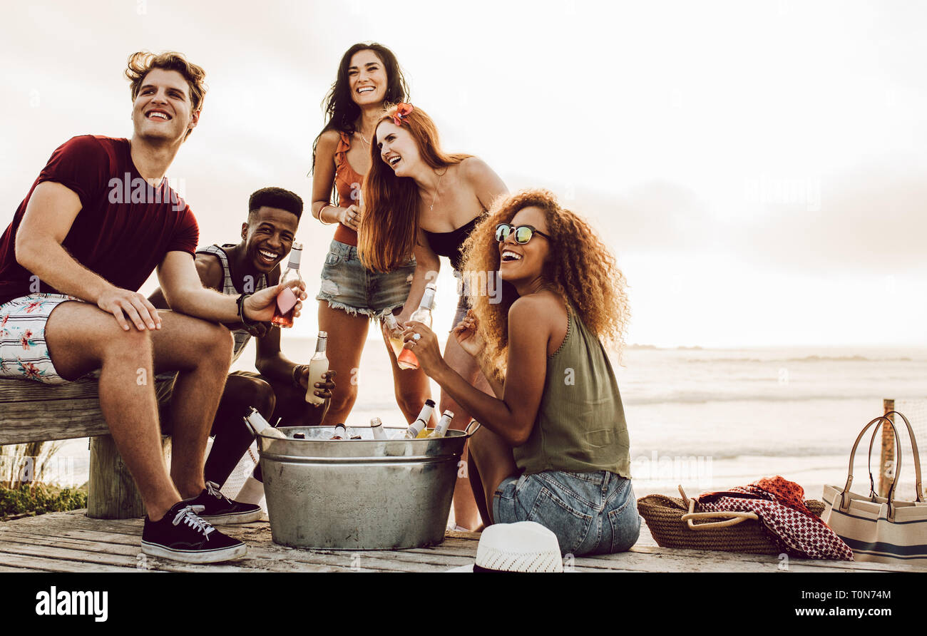 Young people sitting at the beach with beers looking at something interesting. Multiracial friends hanging out at the beach. Stock Photo