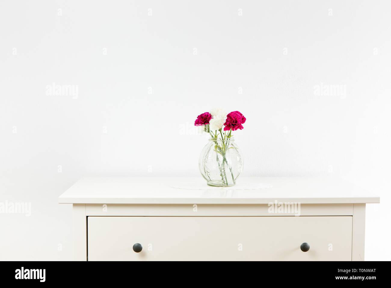 Empty Commode Empty white commode with three flowers on tablecloth. White wall with ...