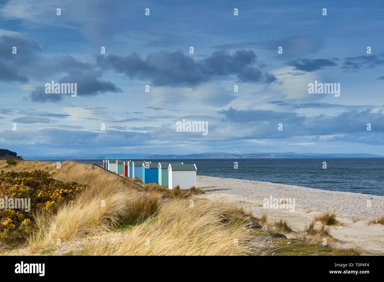 FINDHORN BEACH MORAY FIRTH SCOTLAND COLOURED CHALETS OR BEACH HUTS ON PEBBLE BEACH SNOW OVER THE HILLS AND BLACK ISLE Stock Photo