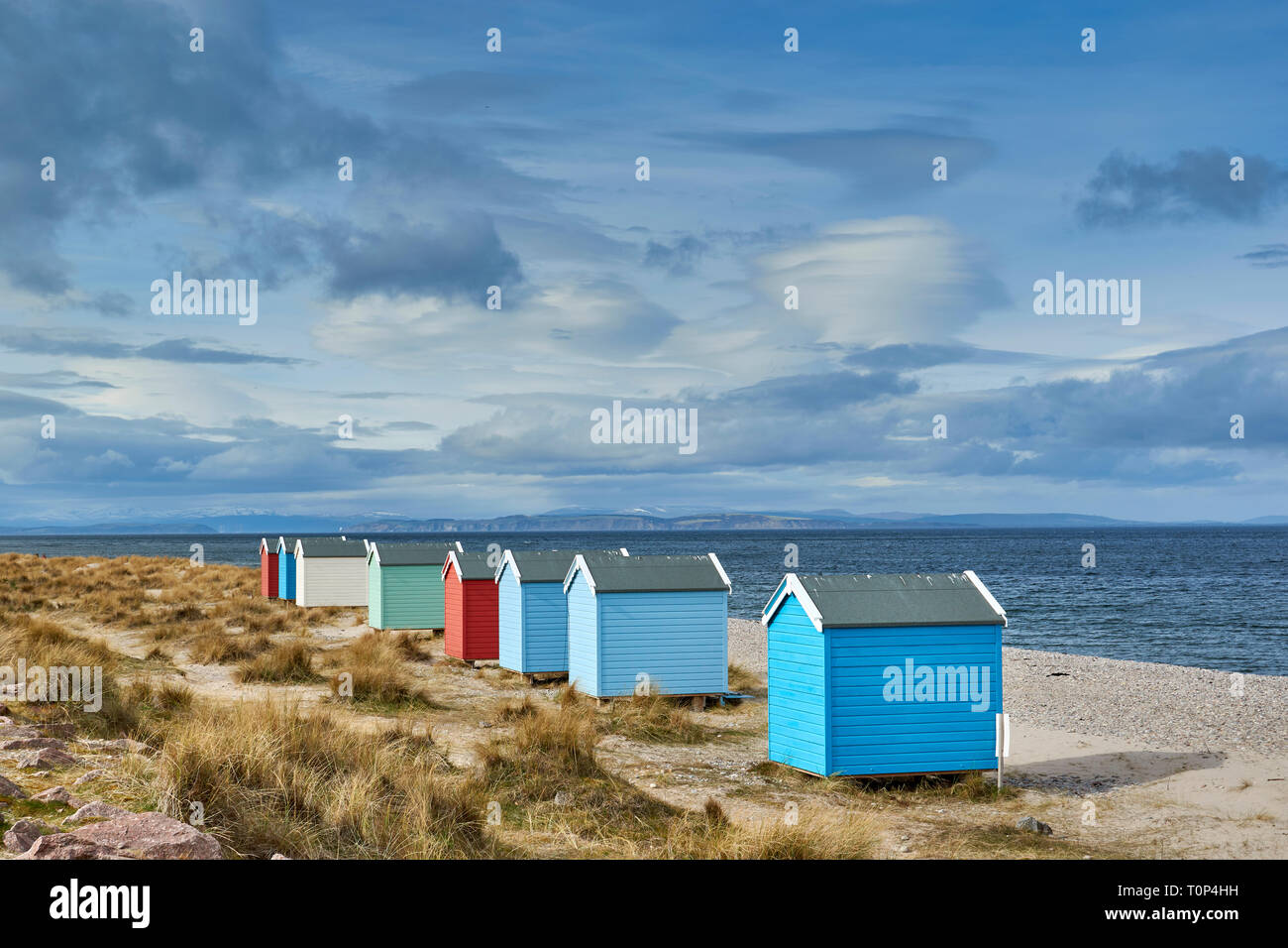 FINDHORN BEACH MORAY FIRTH SCOTLAND EIGHT PASTEL COLOURED CHALETS OR BEACH HUTS ON PEBBLE BEACH SNOW OVER THE HILLS AND BLACK ISLE Stock Photo