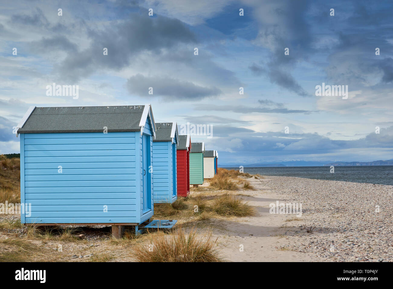 FINDHORN BEACH MORAY FIRTH SCOTLAND PASTEL COLOURED CHALETS OR BEACH HUTS ON PEBBLE BEACH WITH SEA GRASS SNOW OVER THE HILLS AND BLACK ISLE Stock Photo