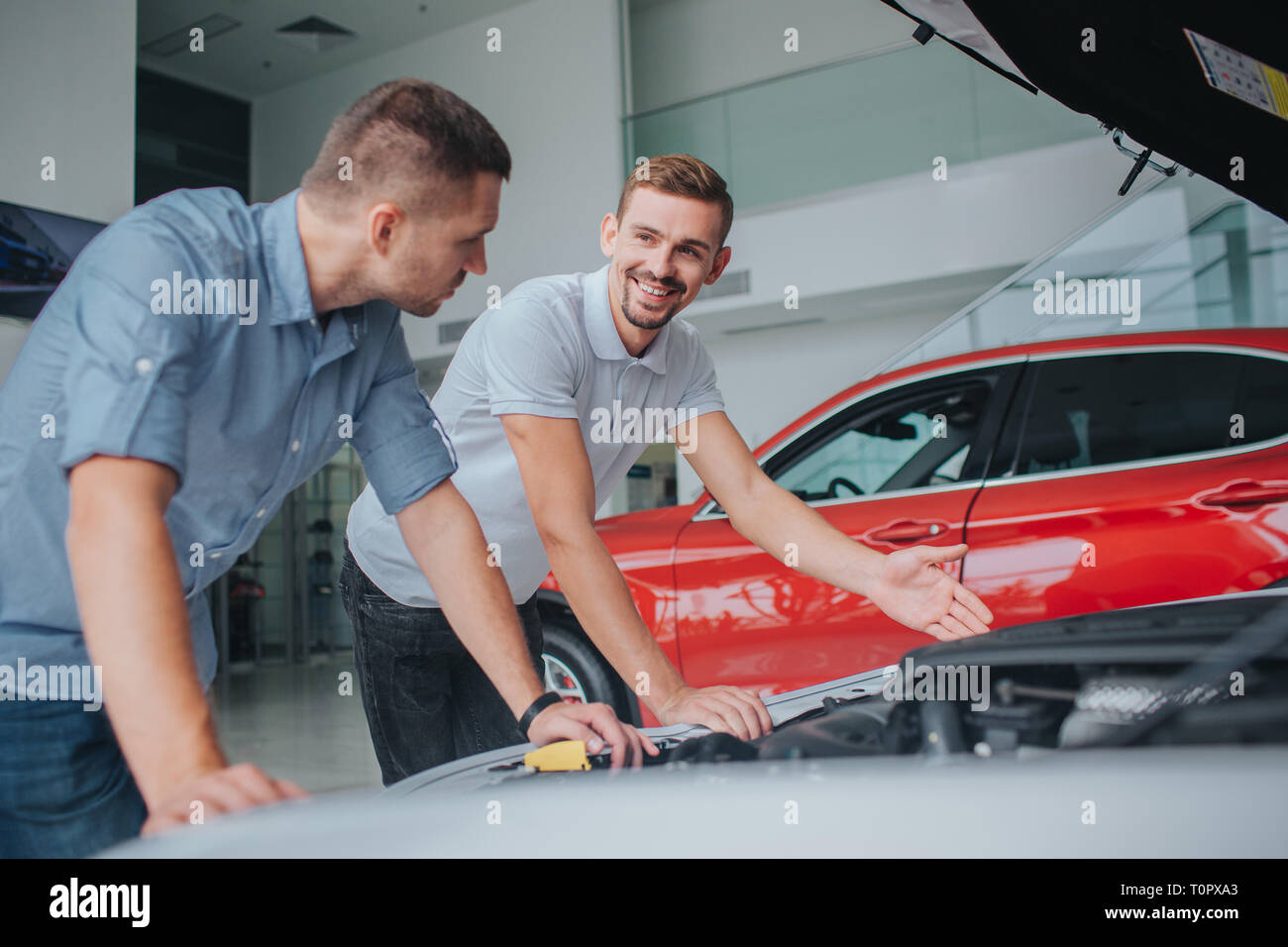 Seller in white sirt stands at opened body of car and point on it. He looks at guy and smiles. Man in grey shirt leans on car and looks at seller. He  Stock Photo
