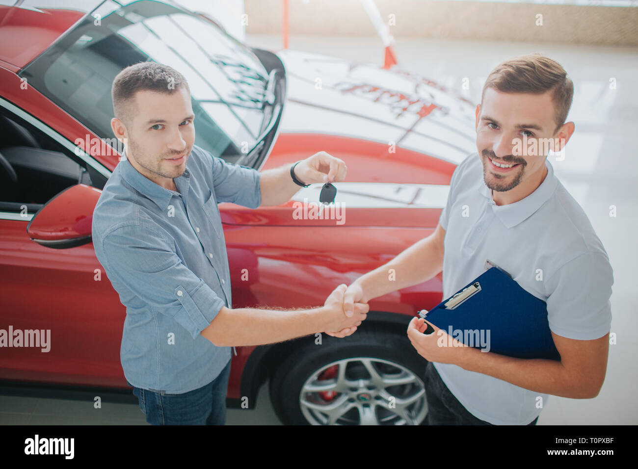 Purchaser and dealer stand and shake each other's hands. Seller holds key from car. Both of look up on camera. They are near red car. Both young men a Stock Photo