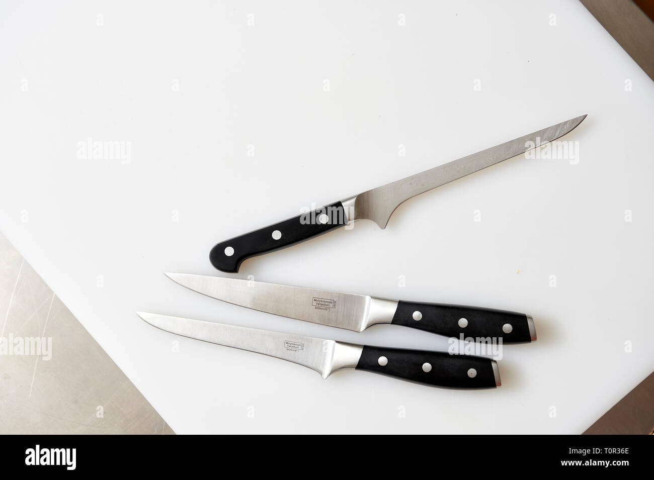 Auf einem weissen Kuechenbrett liegen 3 unterschiedliche Messer fuer die verschiedenen Verarbeitungsschritte in der Kueche. Stock Photo