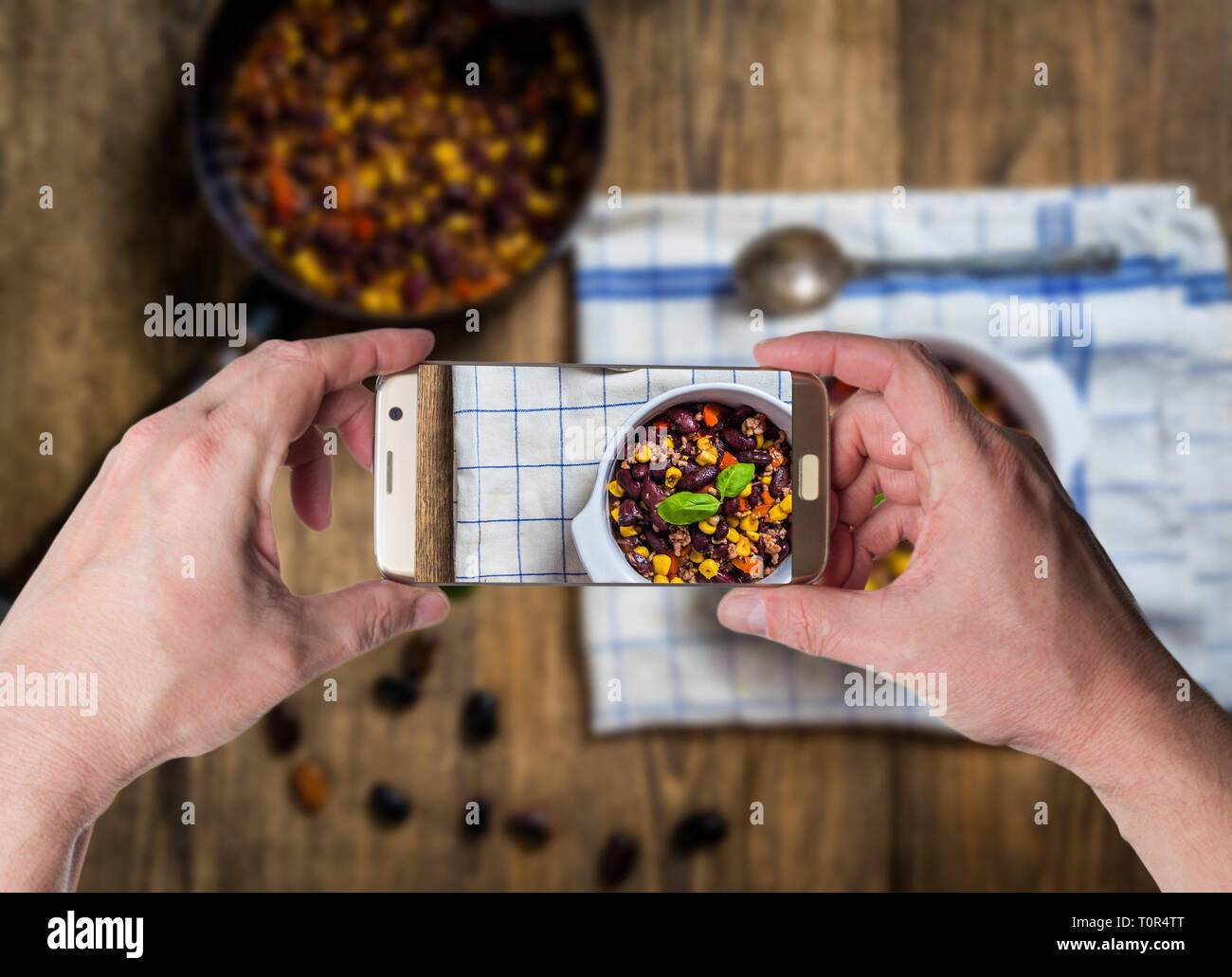 Man taking photo by smartphone of Chili or chilli corn carne. Cooked kidney bean, minced meat, chili, corn and pepper in white bowl  on wood table top Stock Photo