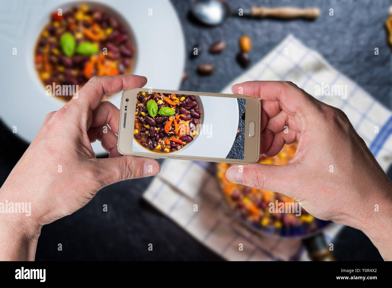 Man taking photo by smartphone of Chili or chilli corn carne. Cooked kidney bean, minced meat, chili, corn and pepper in white bowl  on black stone  t Stock Photo