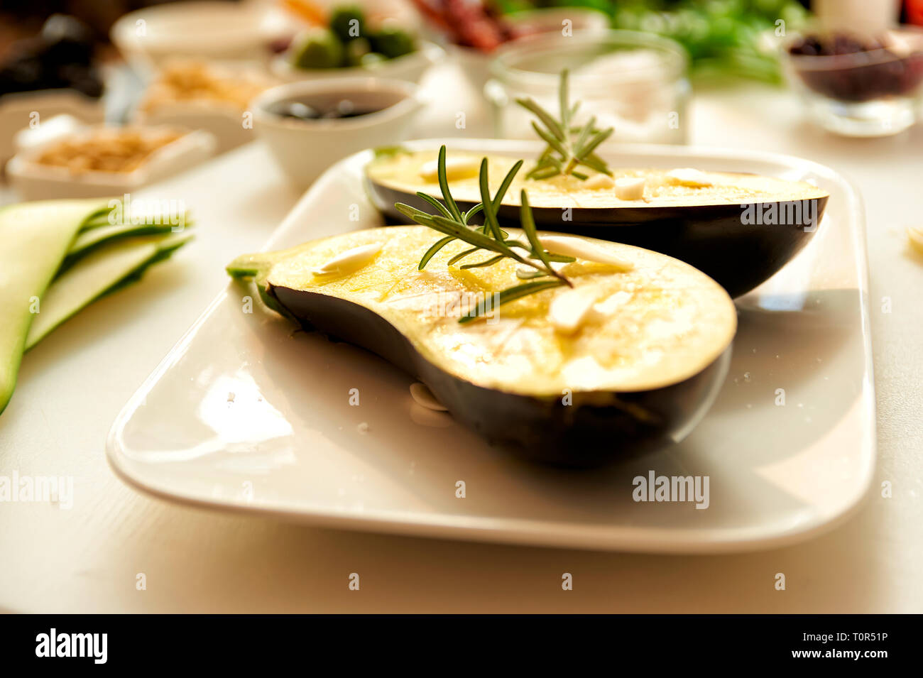 Eine zum Grillen vorbereitete Aubergine die eingeoelt wunde und mit einem frischen Rosmarinzweig belegtwurde liegt auf einer weissen Porzelanschale Stock Photo