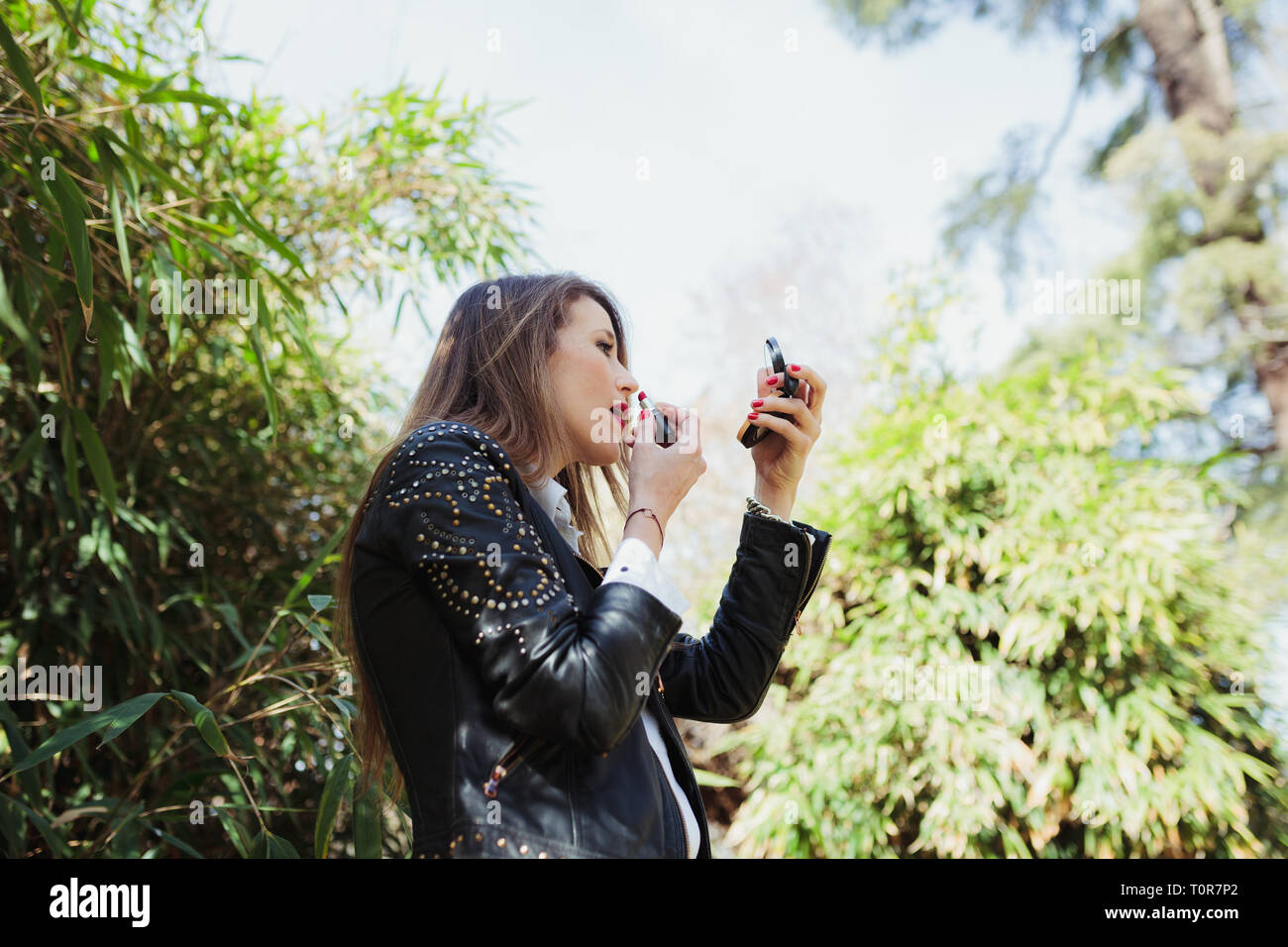 Young blonde woman wearing a leather jacket makes up in the park Stock Photo