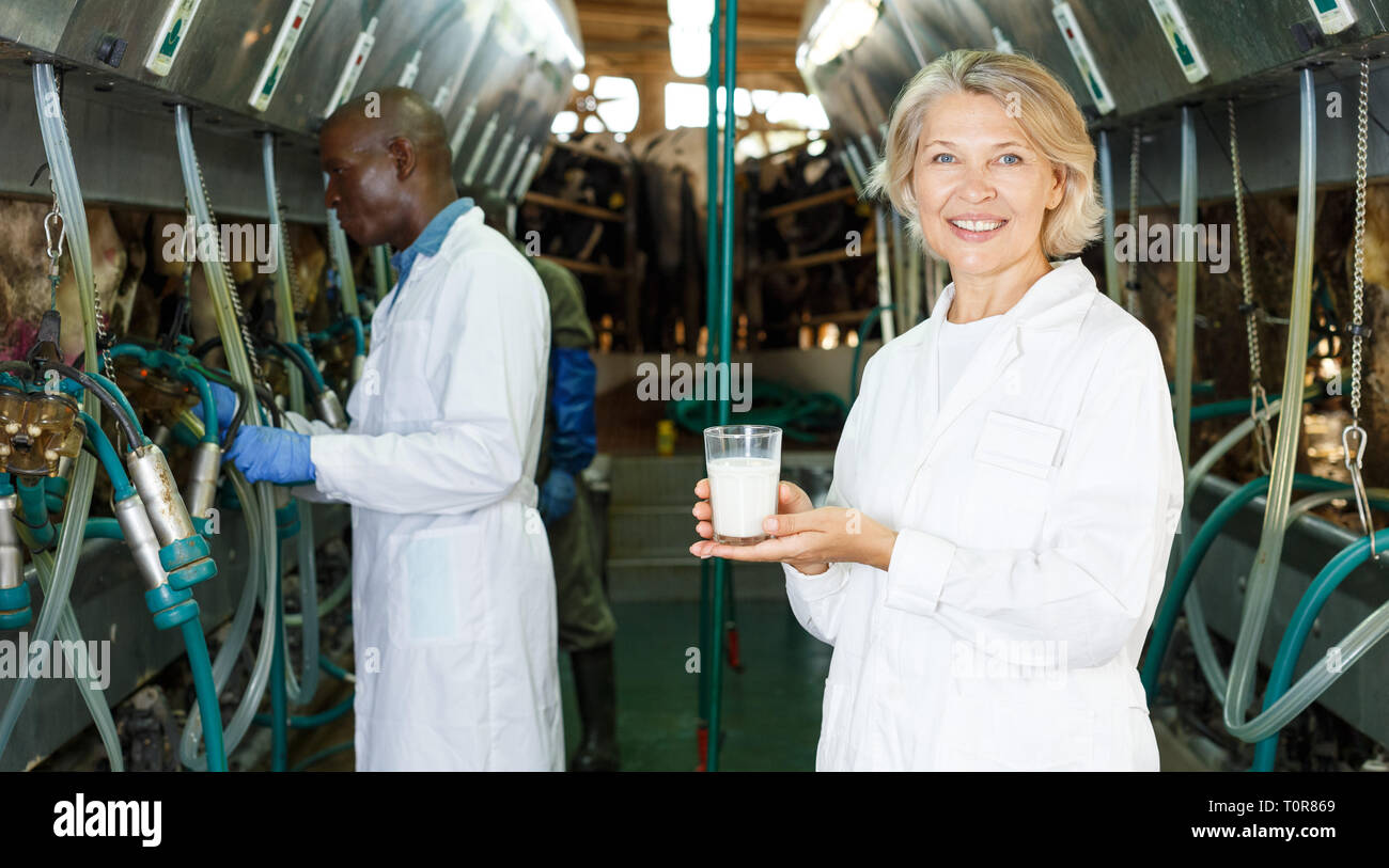 Female dairy engineer in white robe standing with glass of milk near