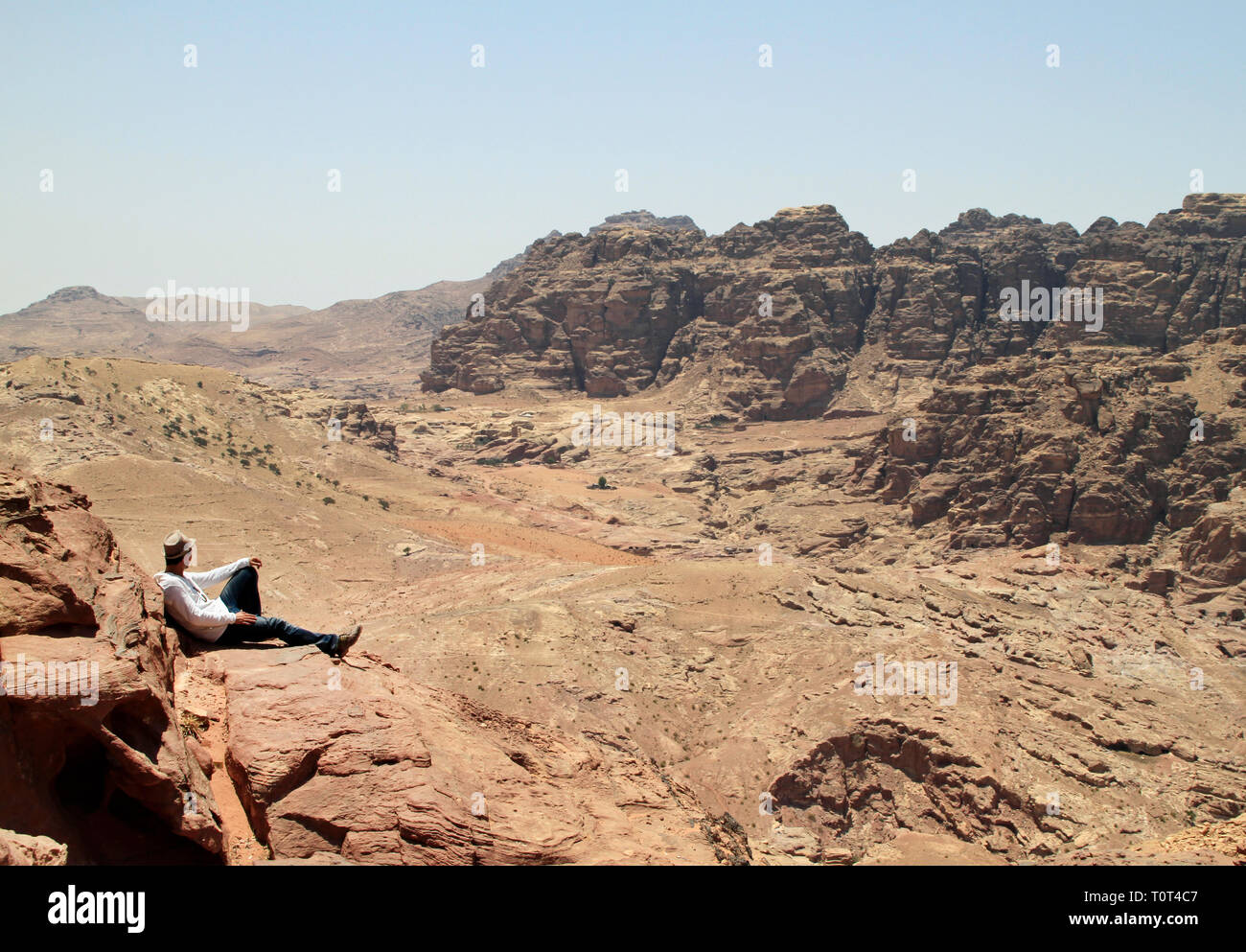 Man wearing a hat sitting on a hill and looking over the rugged landscape of Petra, Jordan Stock Photo