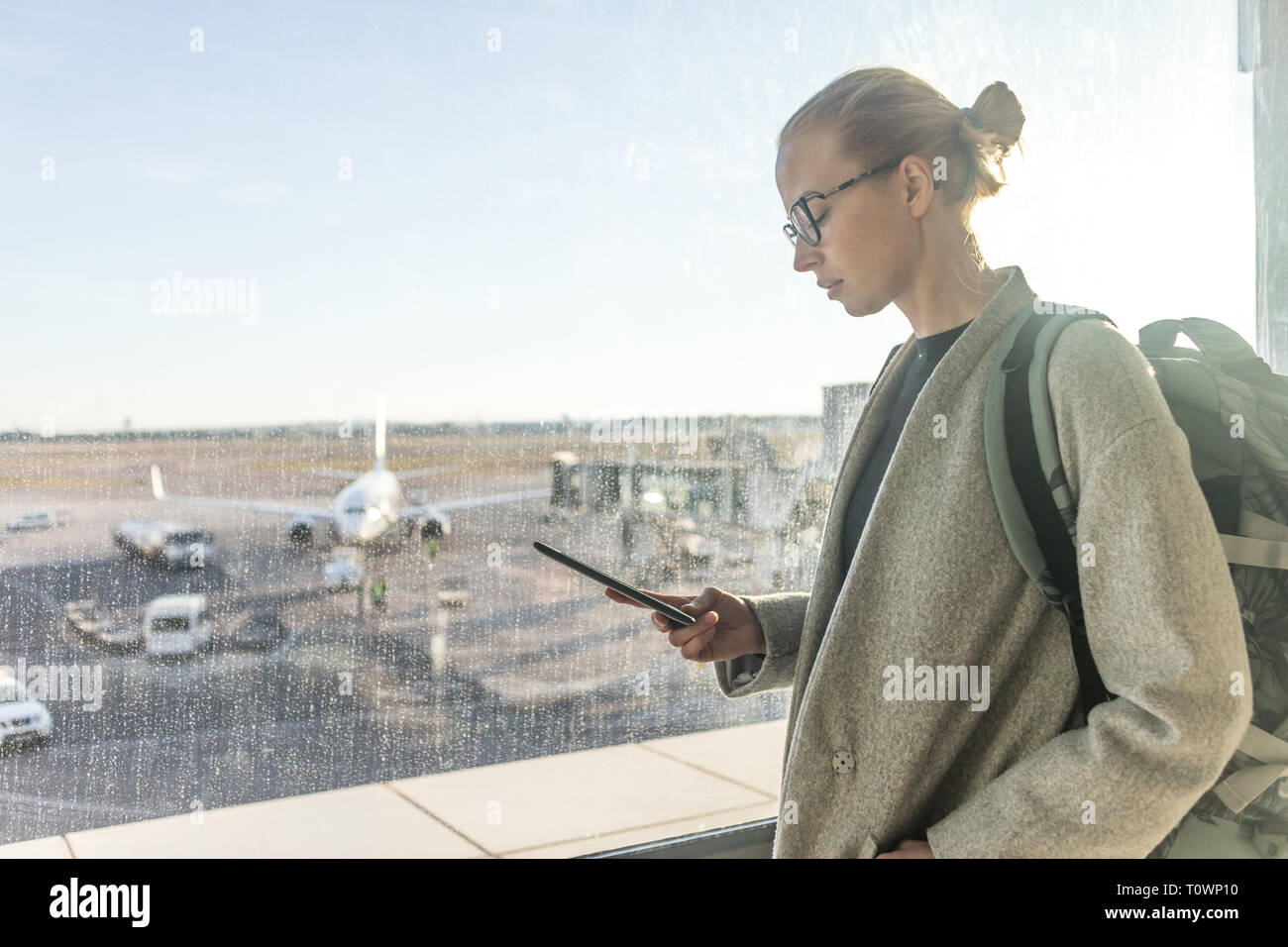 Casually dressed female traveler at airport looking at smart phone device in front of airport gate windows overlooking planes on airport runway Stock Photo