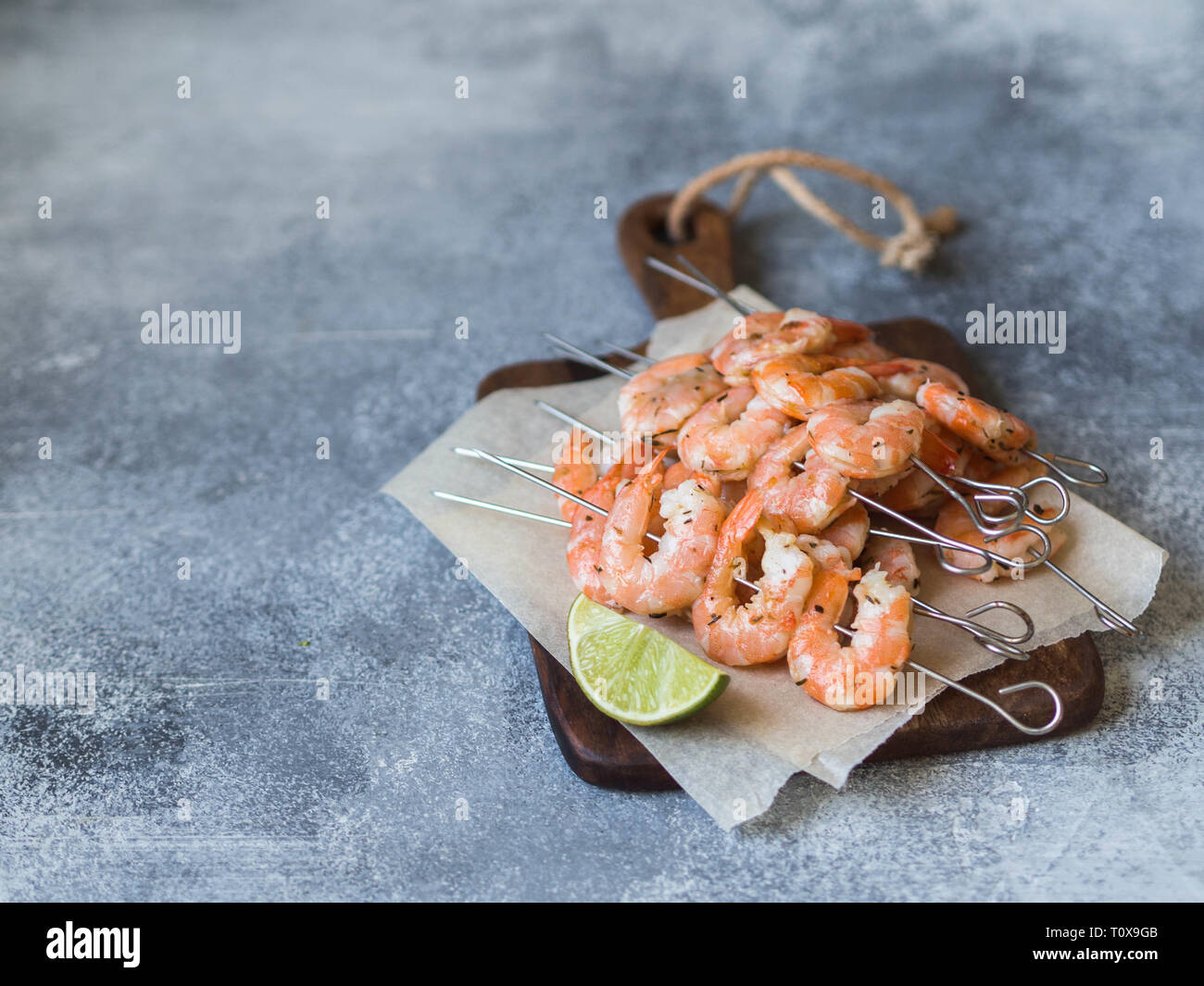 Red shrimps on metal skewers with lime juice on wood board on grey background. Stock Photo