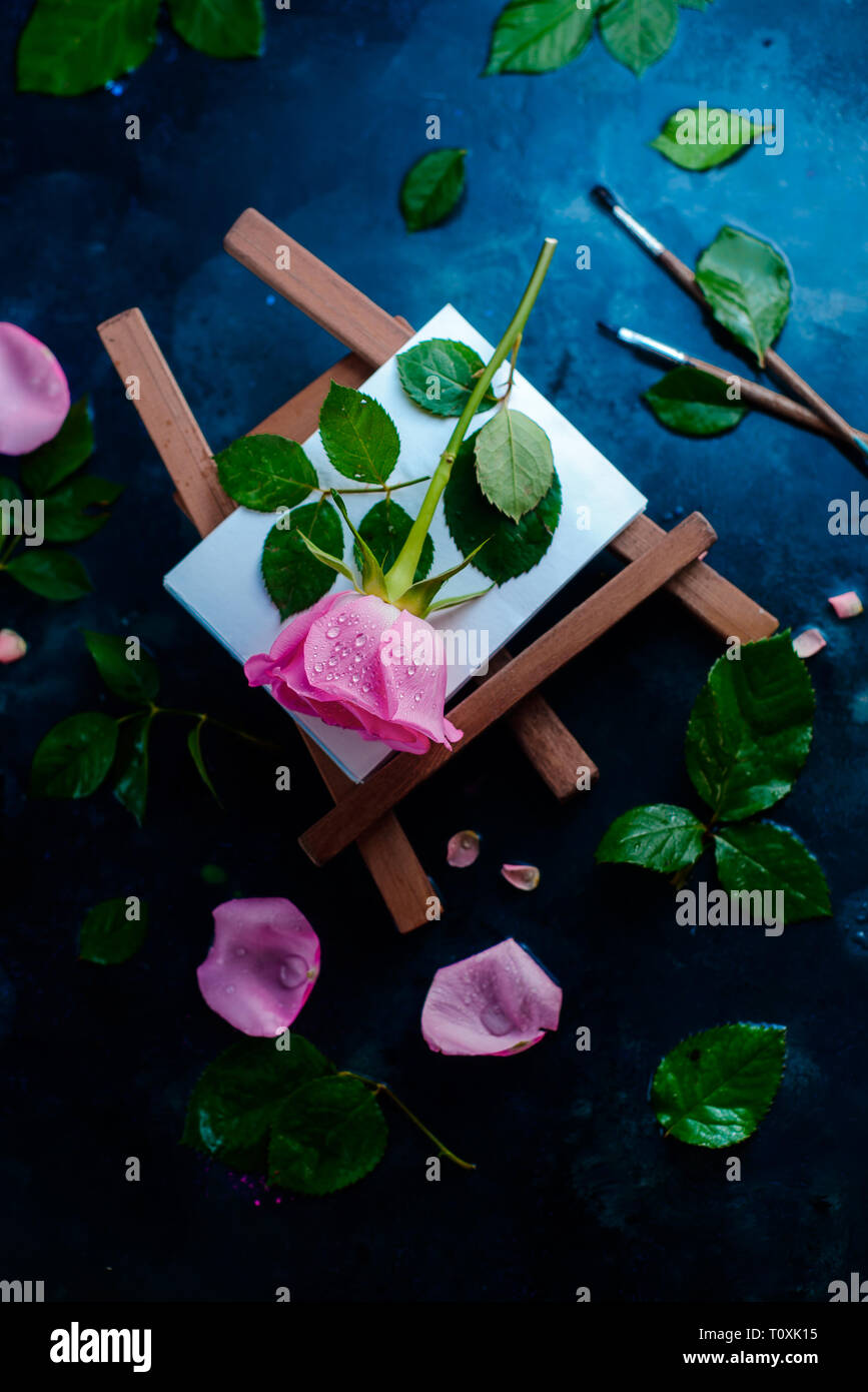 Rose flower on a painter easel flat lay. Painting tools, brushes and pencils on a wet rainy background with copy space. Nature and art concept Stock Photo