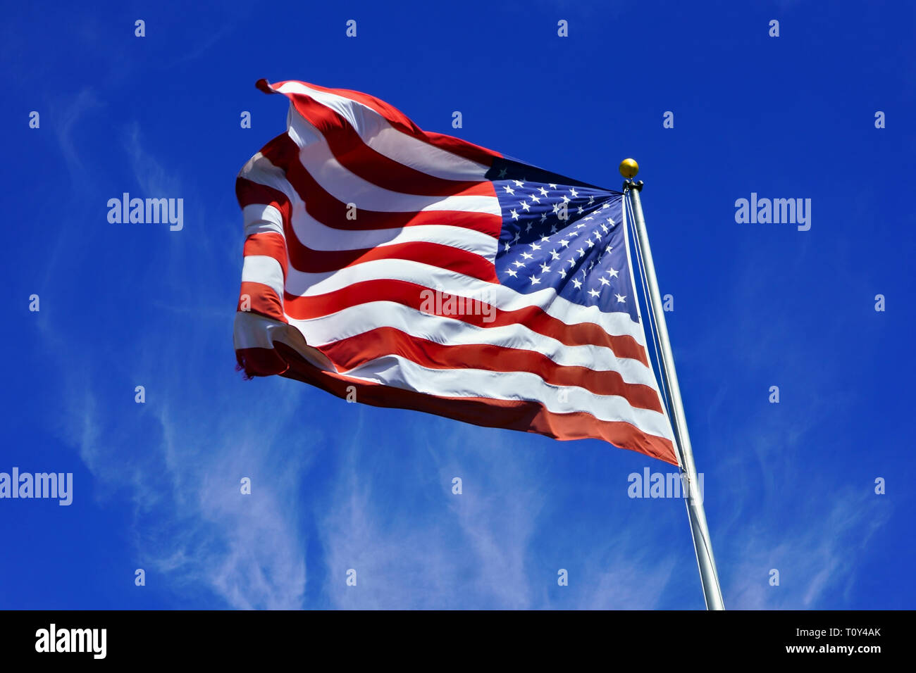 US flag, dynamically fluttering in the wind against a blue cloudy sky background. American flag with stars and stripes in strong colors. Stock Photo