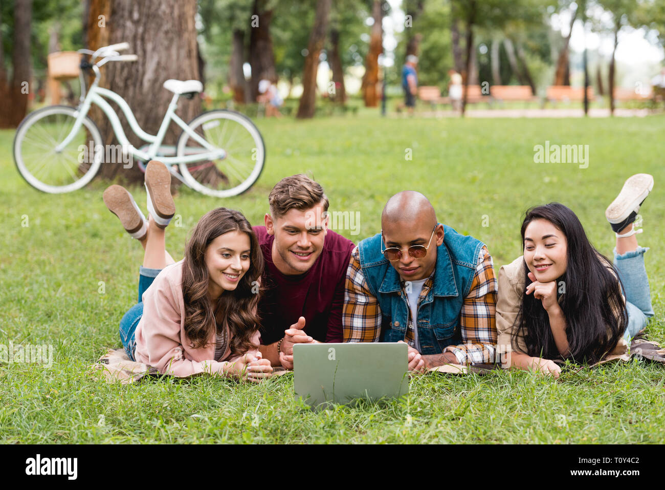 happy multicultural young men lying with beautiful girls on blanket and looking at laptop Stock Photo