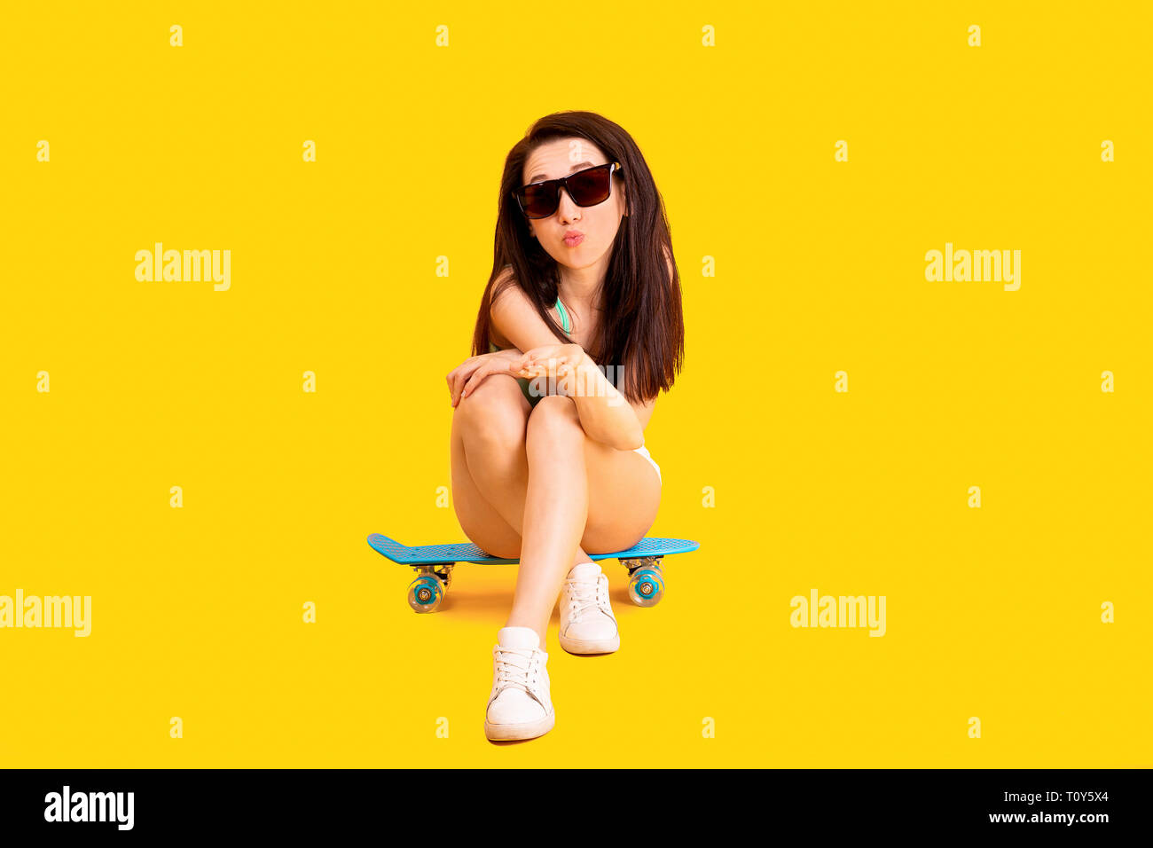 beautiful girl in sunglasses, sitting on a skateboard and sending air kiss, image on yellow background Stock Photo
