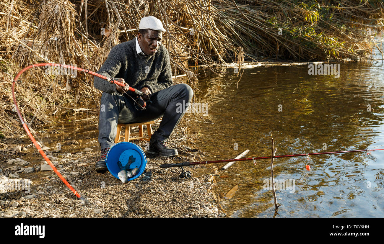 Portrait of afro fisherman sitting on stool and pulling fish Stock Photo