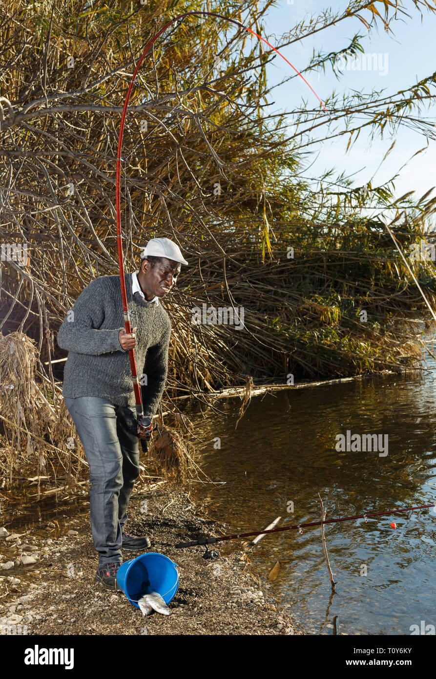 Mature African man standing near river and pulling fish expressing emotions of dedication Stock Photo
