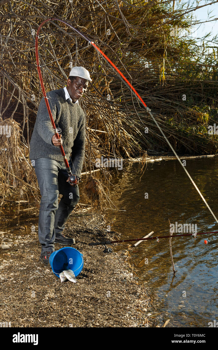 Mature African man standing near river and pulling fish expressing emotions of dedication Stock Photo