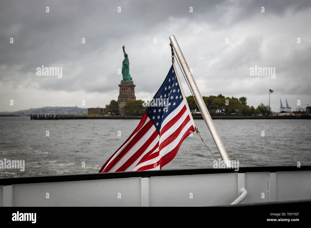 An American flag waves on the back of a boat leaving the Statue of Liberty visible behind on a cloudy, rainy day in September in New York City. Stock Photo