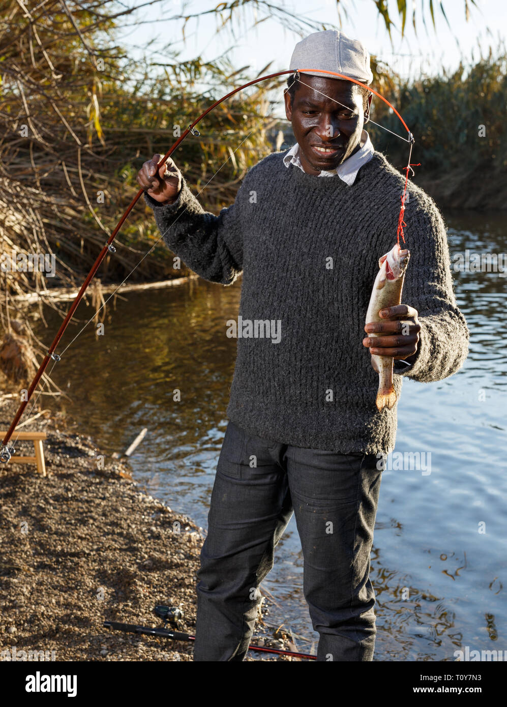 Portrait of enthusiastic African man holding fishing rod with fish on hook Stock Photo