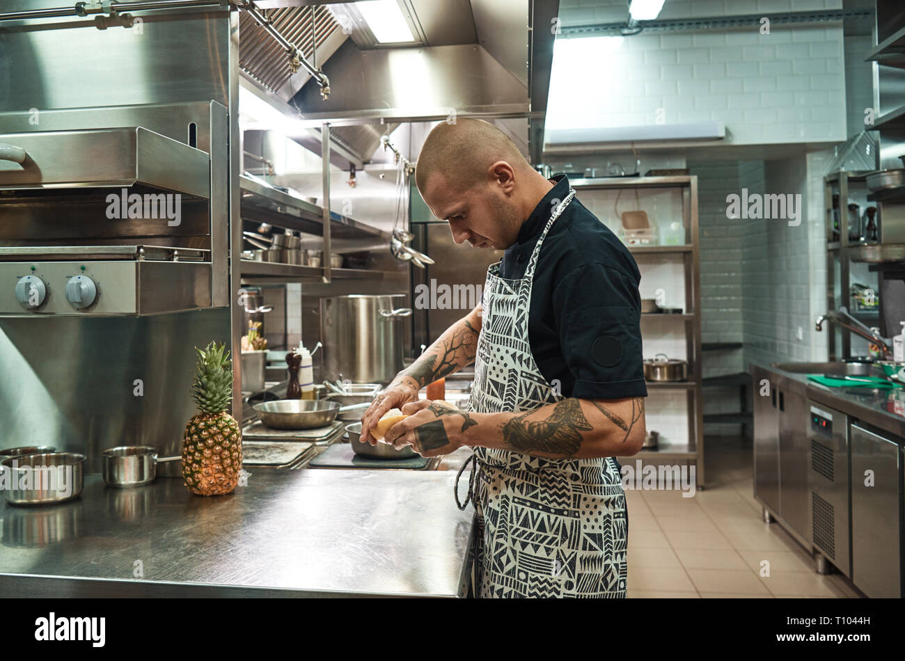 Fresh fruits. Side view of young chef with several tattoos on his arms peeling a grapefruit while standing in a professional kitchen. Making a juice Stock Photo