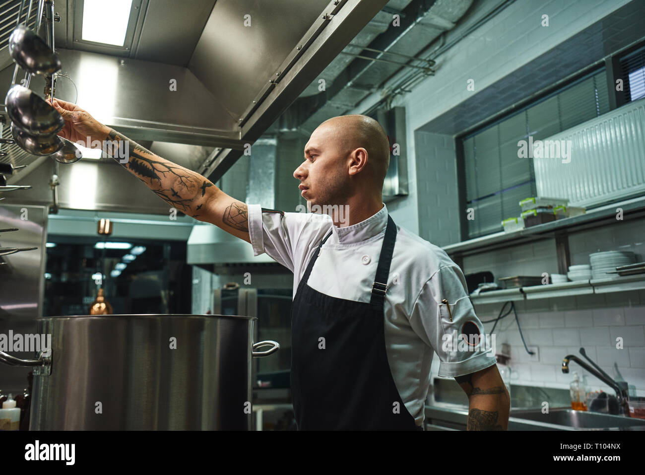 Famous soup. SIde view of attractive bald chef in apron taking a ladle for soup cooking while standing in a restaurant kitchen. Cooking concept Stock Photo