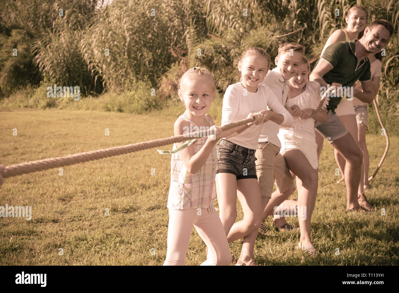 Positive kids with moms and dads playing tug of war during joint outdoors games on sunny day Stock Photo