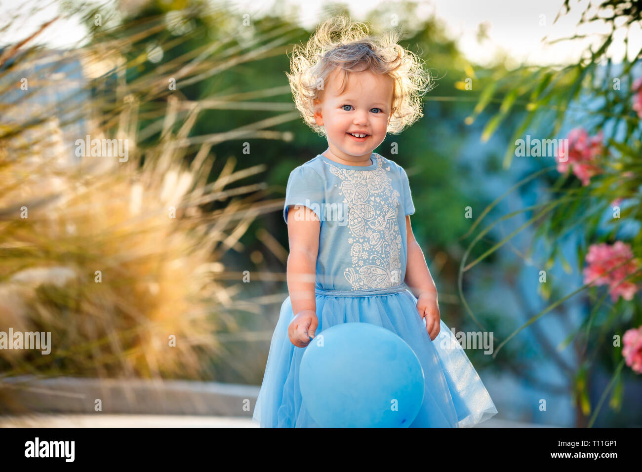Portrait of happy little girl with curly blonde hair in casual clothes posing outdoors with blue ballons on hand Stock Photo