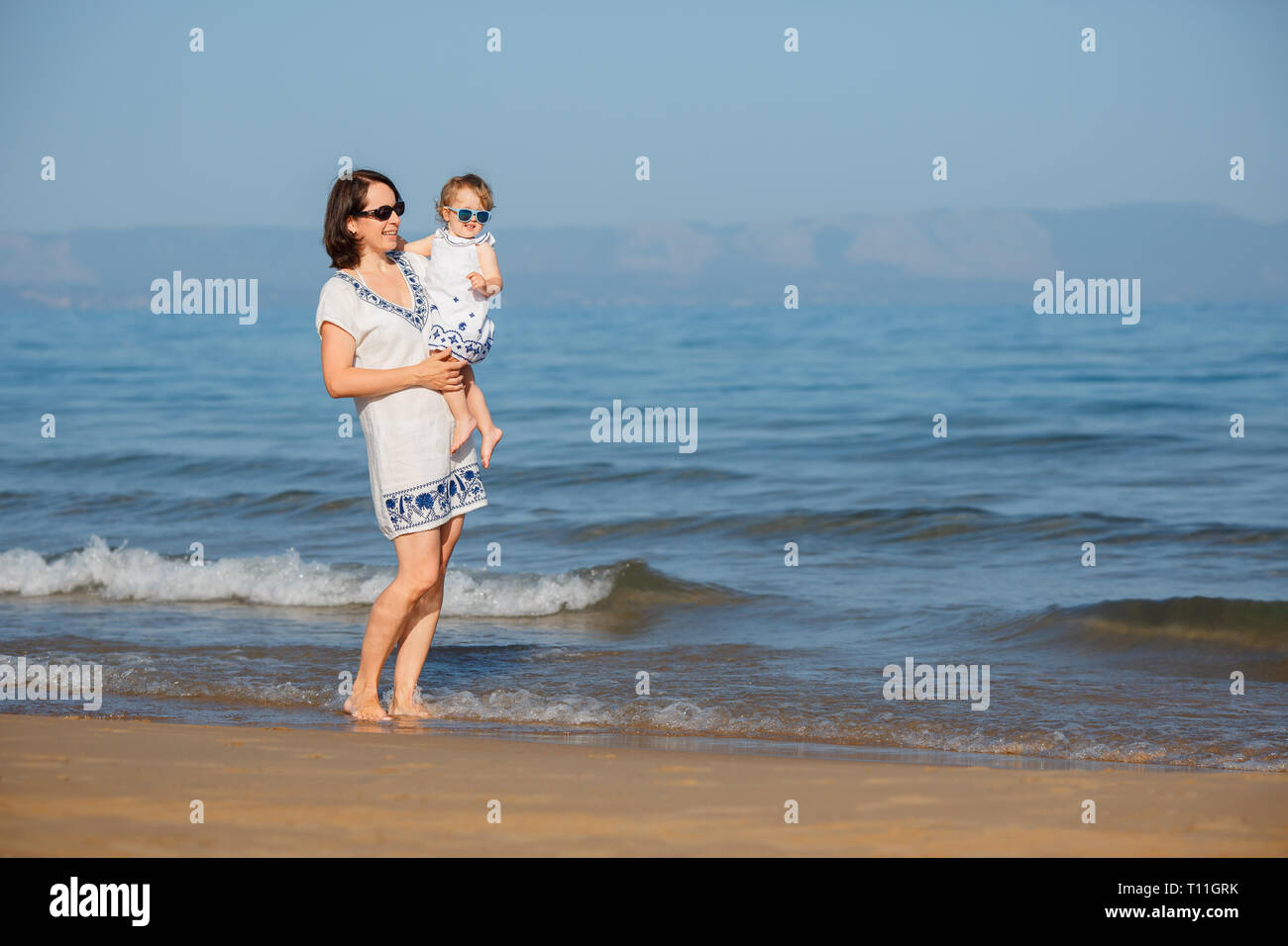 Young mother and her cute little baby girl playing on a beautiful tropical beach enjoying family summer holiday. Family sea summer vacation. Ocean and Stock Photo