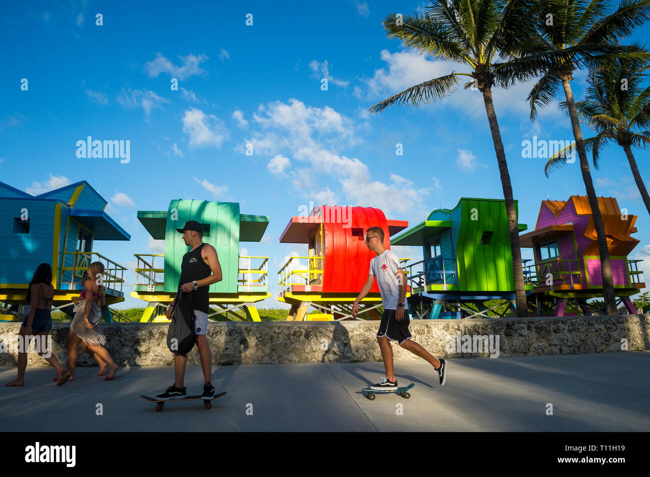 MIAMI - JULY 2017: Young men on skateboards pass along the Miami Beach promenade in front of vibrantly colored lifeguard towers in Lummus Park. Stock Photo