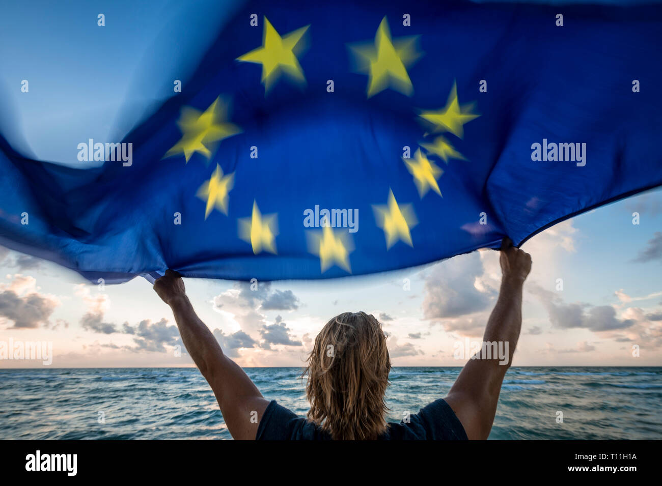 Scenic sunny view of man with blond hair holding a European Union flag waving in strong motion blur in the wind in front of a soft sunrise beach scene Stock Photo