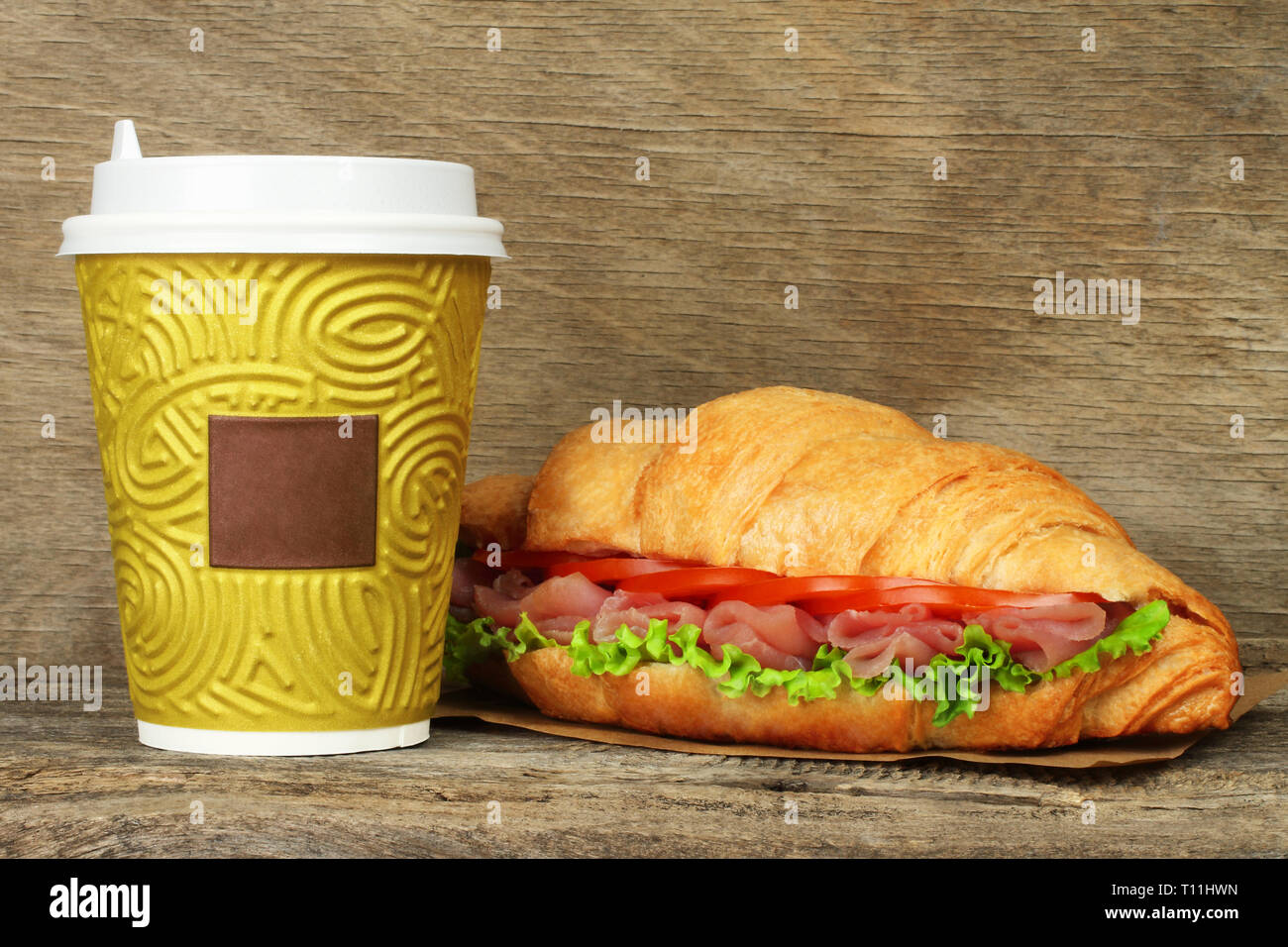 Big croissant with green salad, tomatoes and pork meat near paper coffee cup on old wooden background Stock Photo