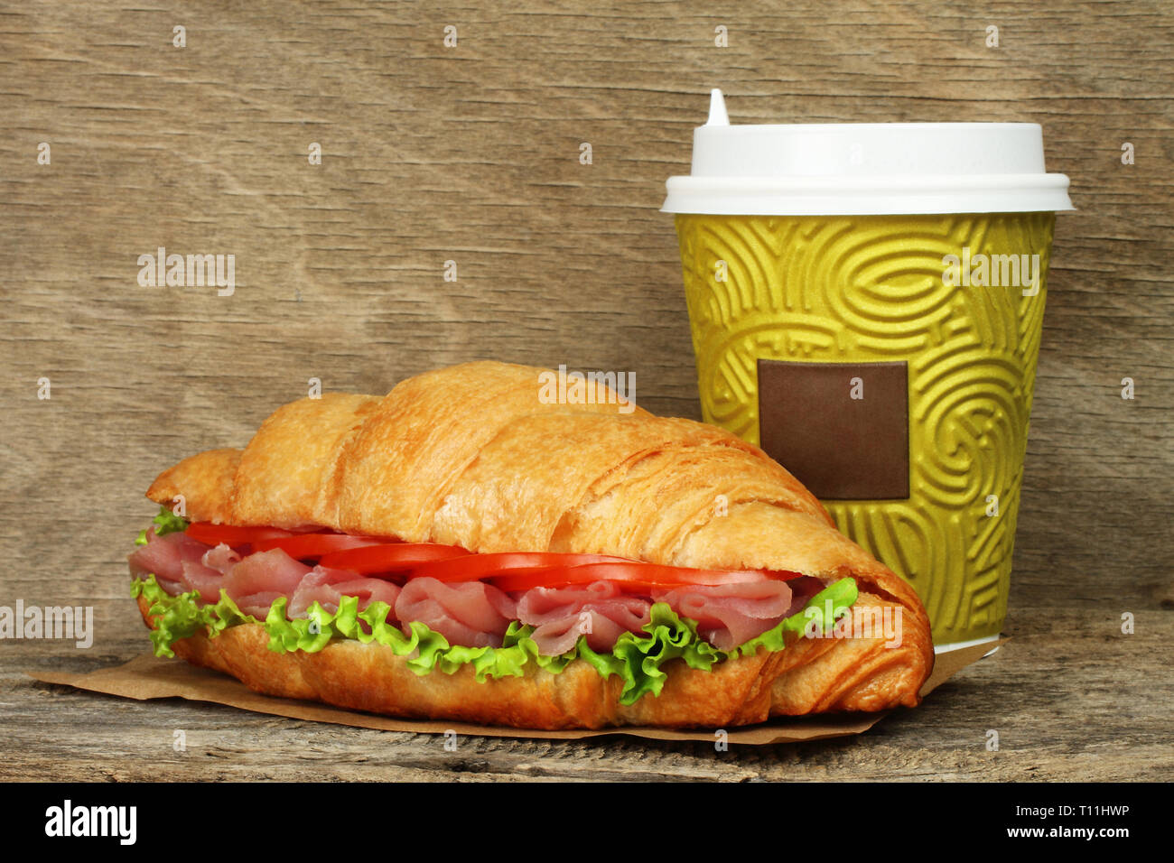 Big croissant with green salad, tomatoes and pork meat near paper coffee cup on old wooden background Stock Photo