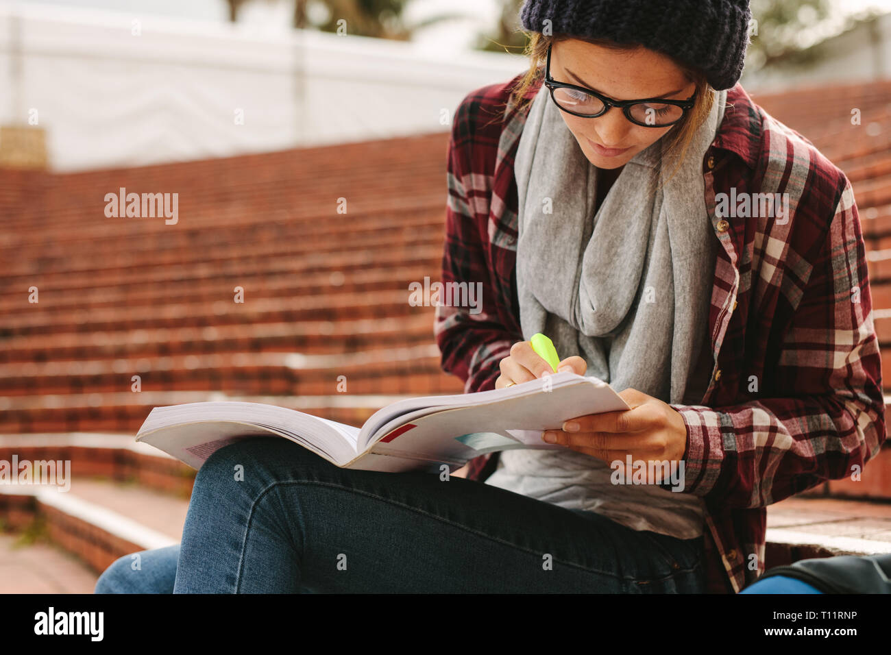 Female student sitting on steps in college campus marking important points in the book with a highlighter pen. University student studying at campus. Stock Photo