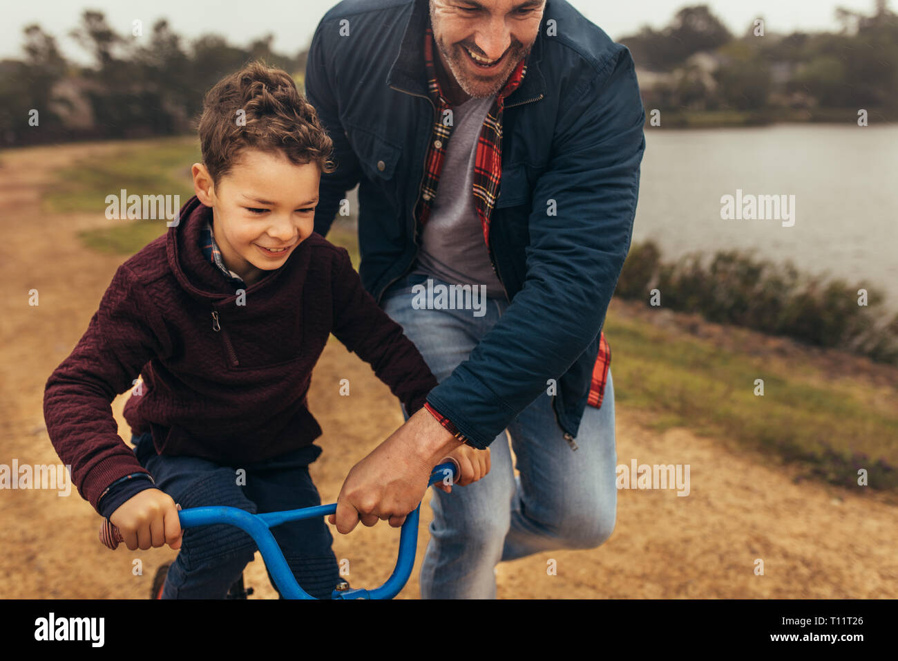 Smiling kid learning to ride a bicycle near a lake. Man walking along the bicycle holding it while the kid learns to ride it. Stock Photo
