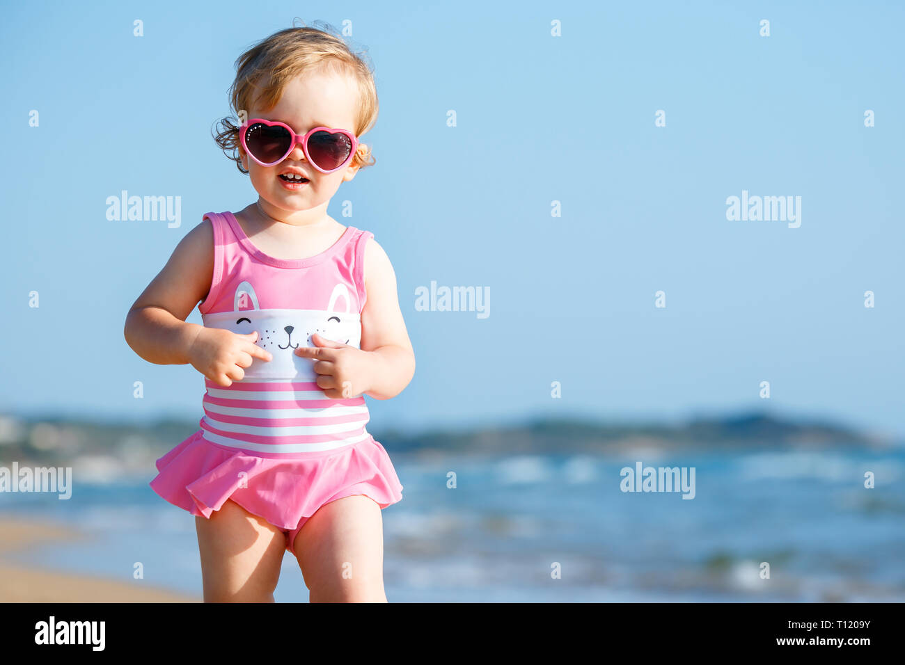 Cute curly baby girl playing on a beautiful tropical beach wearing a cute swimsuit and sunglasses Stock Photo