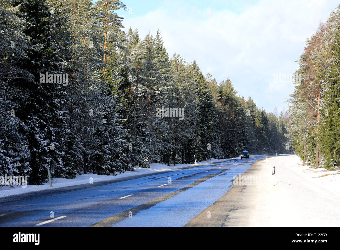 Landscape of a wet, rural highway flanked by forest with car approaching in the distance on a sunny day of winter. Marttila, Finland. Stock Photo