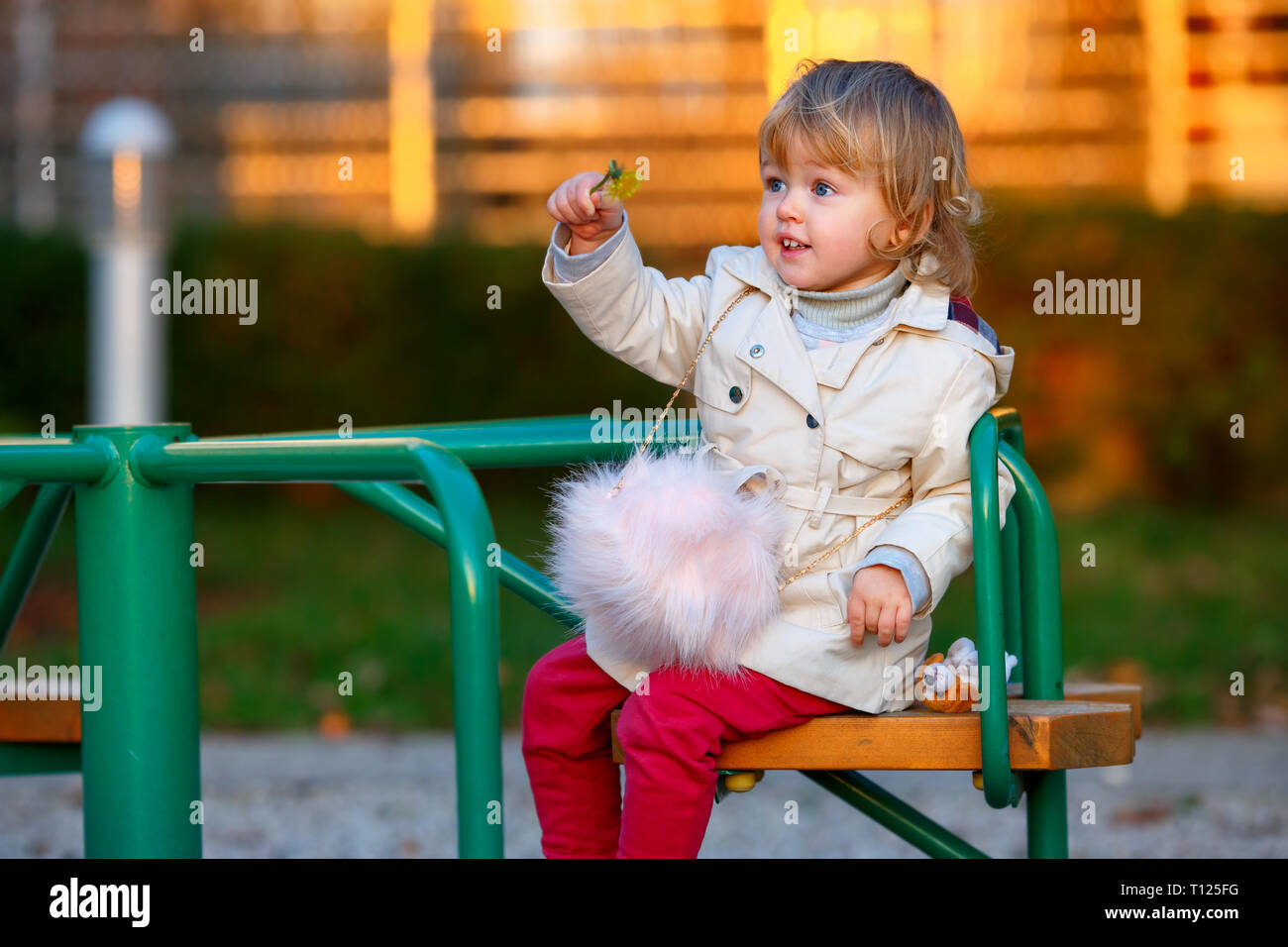 Portrait of sweet blond little baby girl in trench coat and faux fur bag at playground Stock Photo