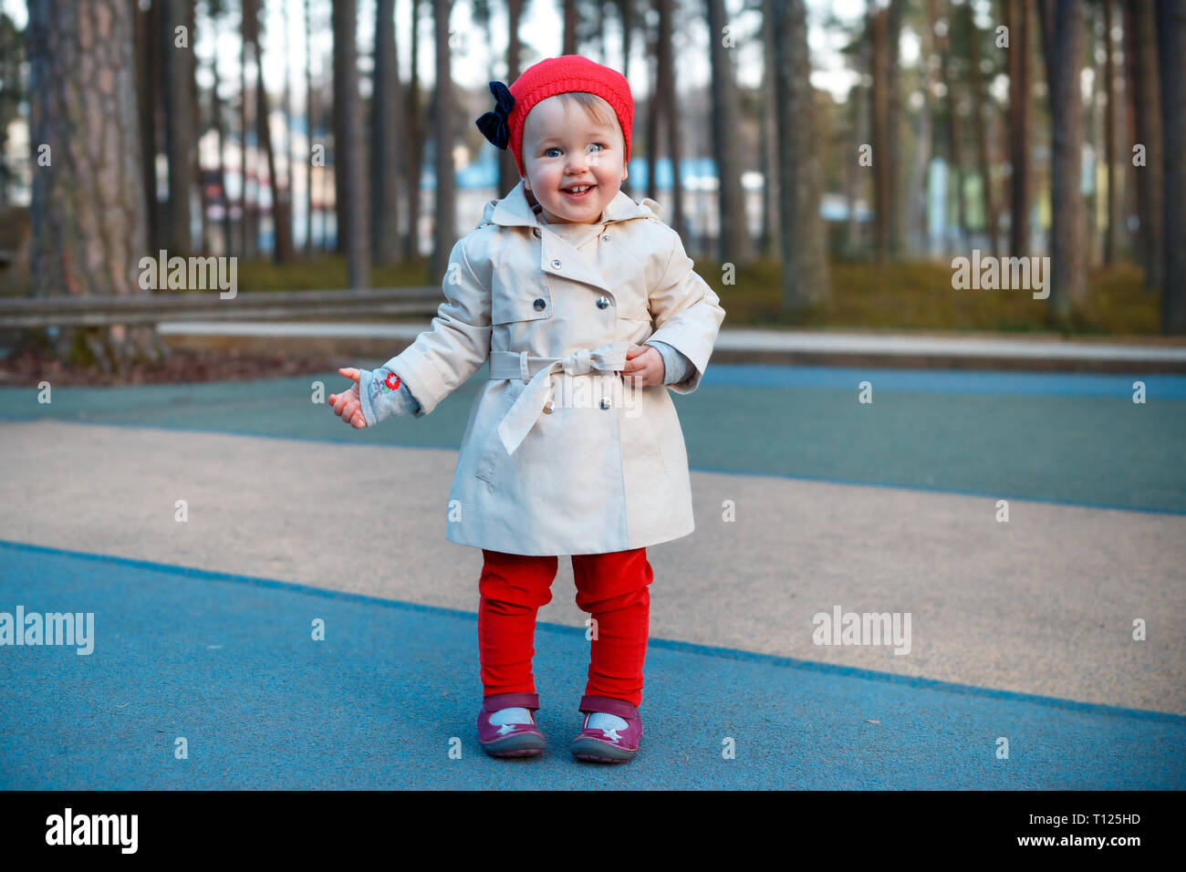 Sweet blond little toddler girl in beautiful beige trench coat and red cap plays outdoors at playground Stock Photo