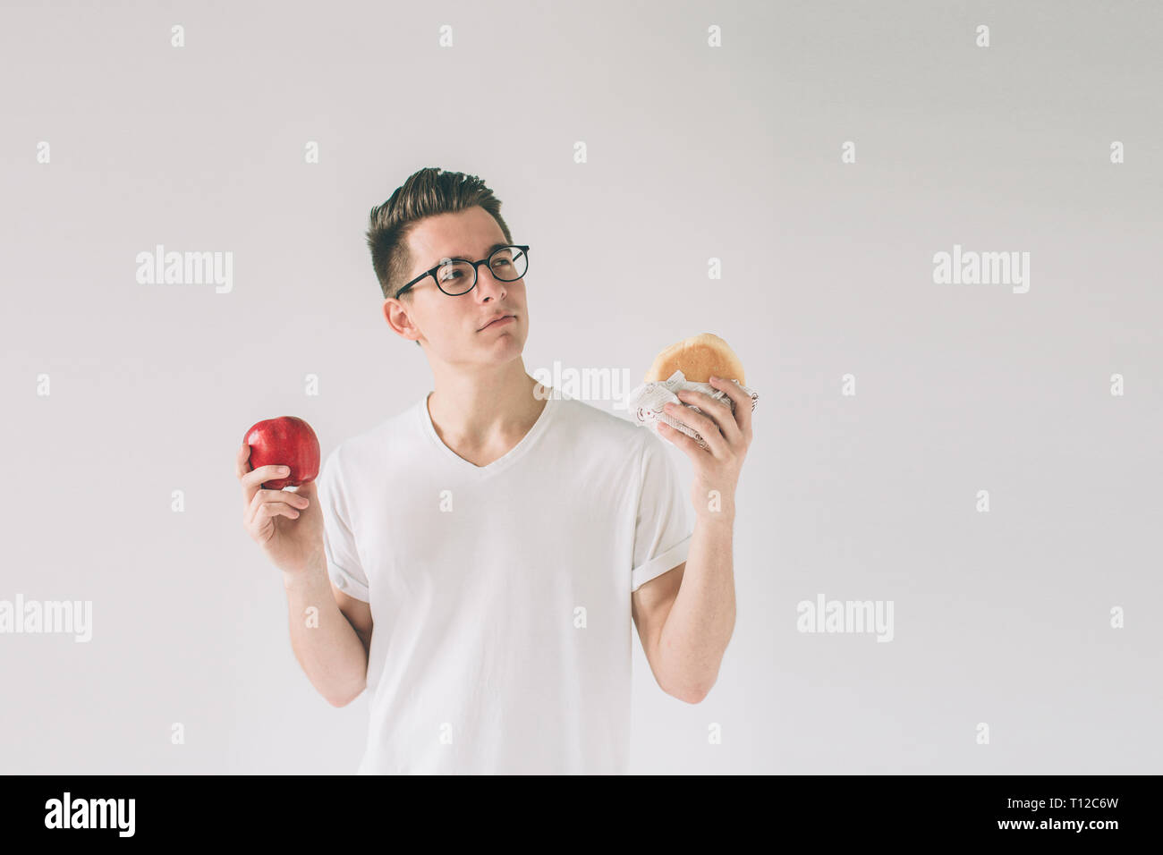 Bearded man in a white shirt on a light background holding a hamburger and an apple Man makes the choice between fast and healthy food. Tasty or usefu Stock Photo