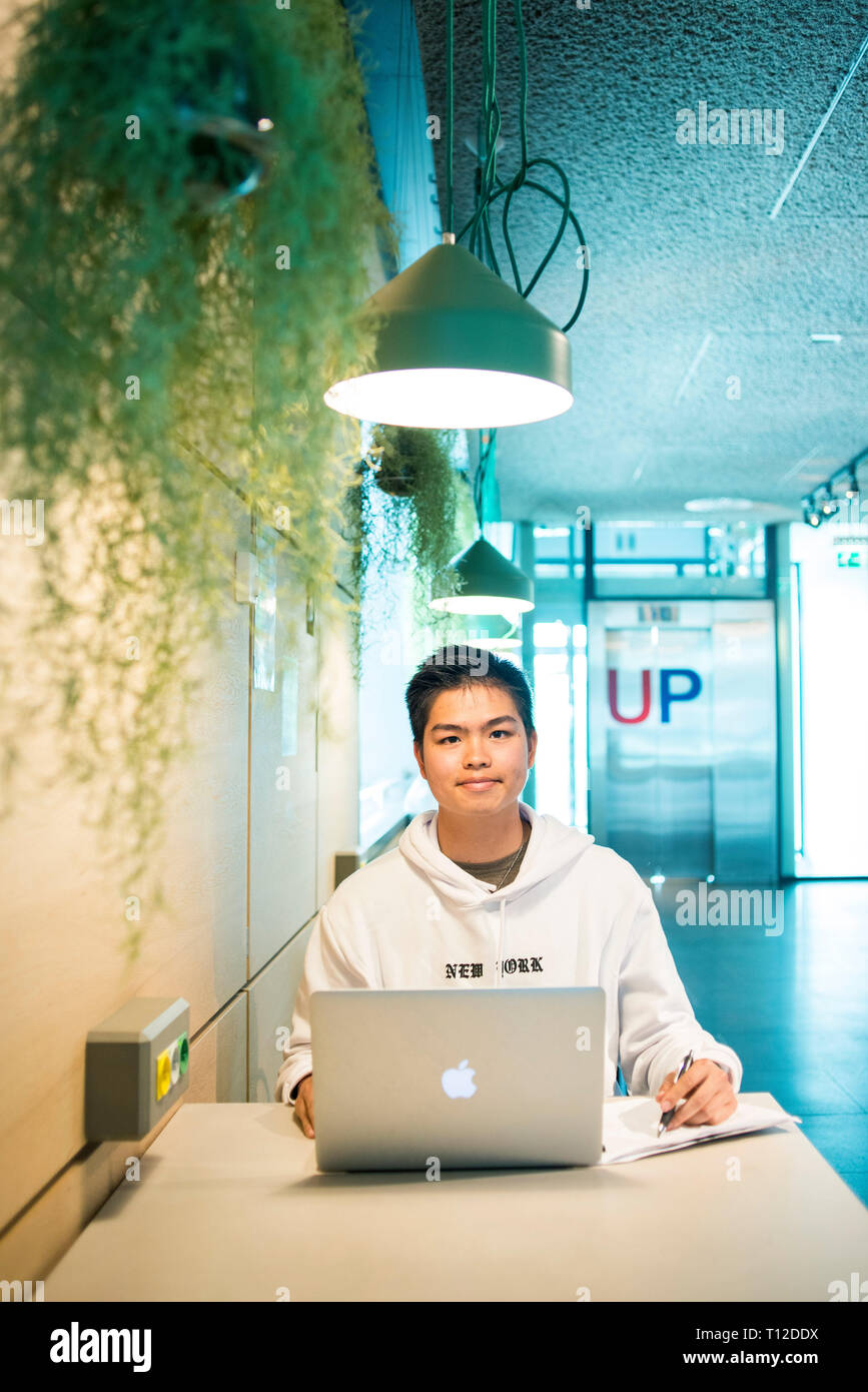 A young Asian male student sits at a table and works on his laptop ...