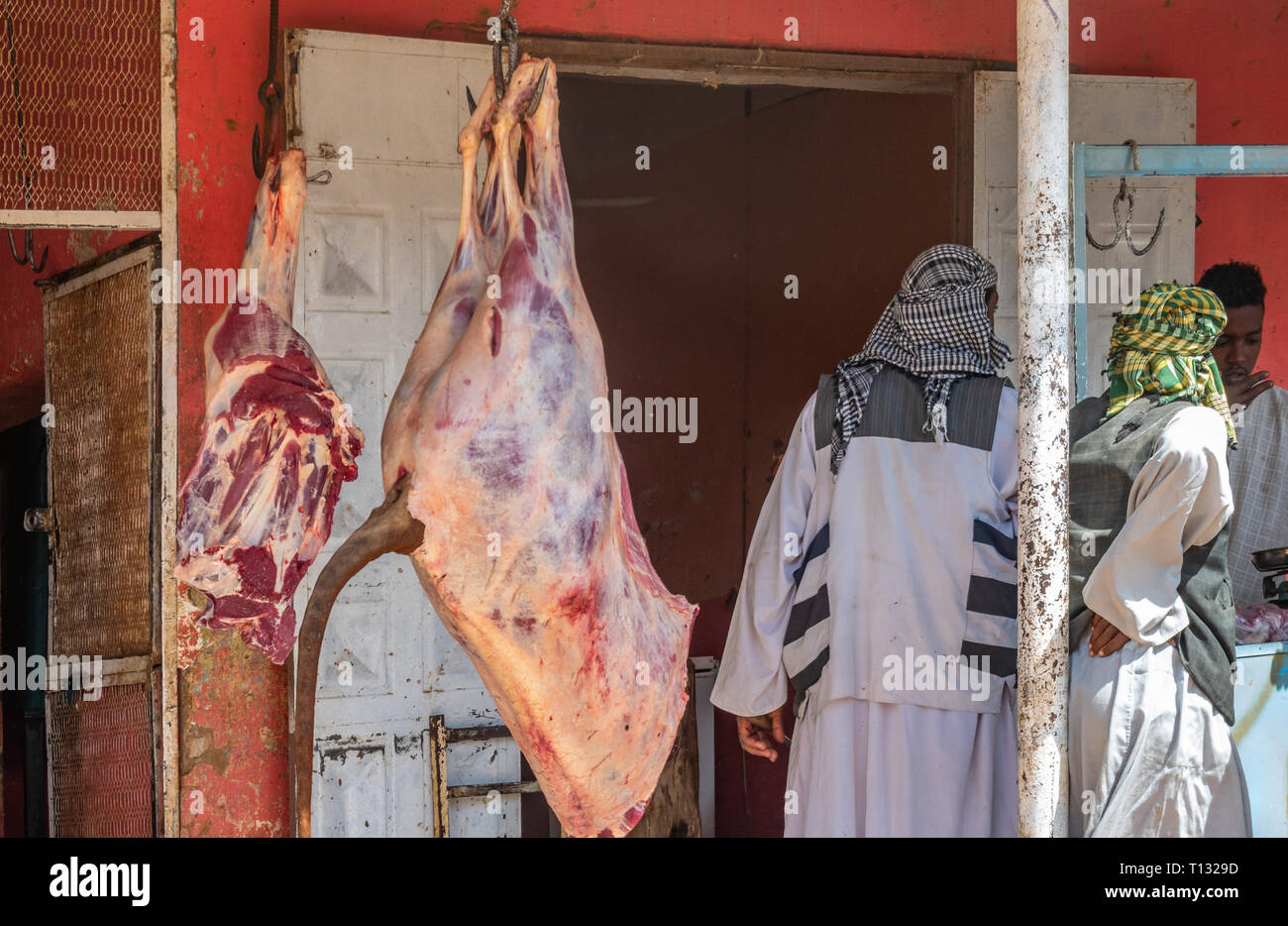 Nuri, Sudan, February 9., 2019: A large piece of beef hangs in front of a butcher's while men negotiate with the butcher in caftans and turban. Stock Photo